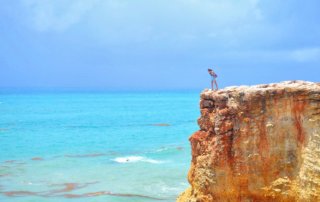 Tiny human standing on tall cliff overlooking light blue ocean water