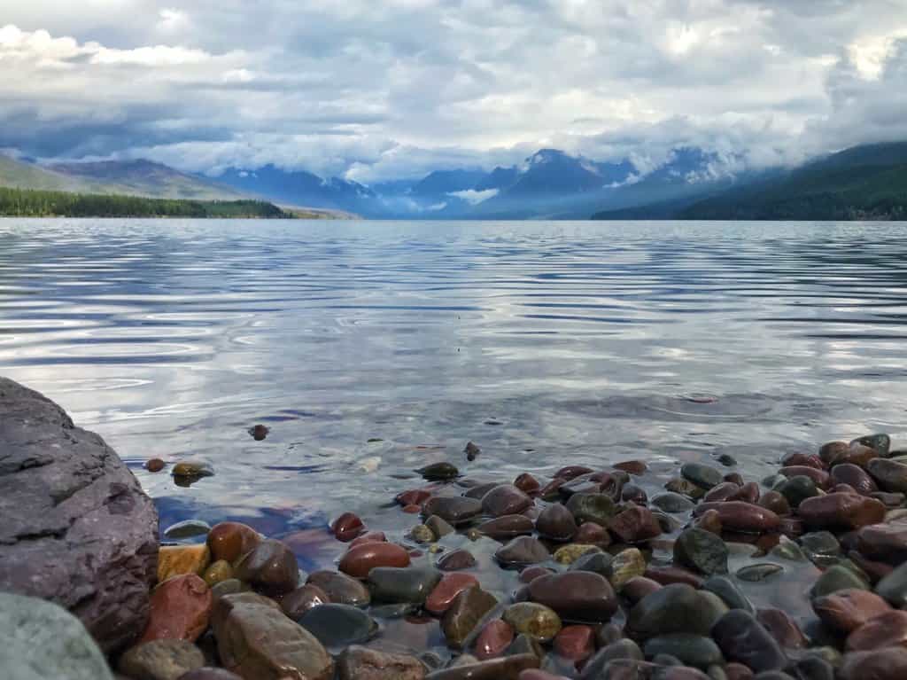 Campgrounds in Glacier National Park near Lake McDonald, colored rocks, clear water, and snow-covered mountaintops in the background